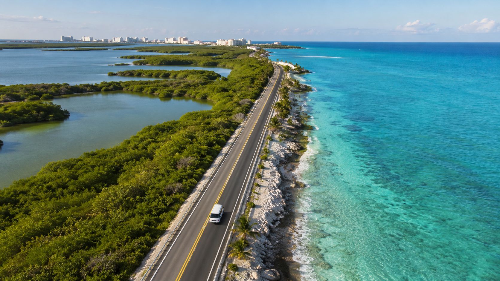 An aerial view of a single road stretching along the coast between mangroves and turquoise ocean waters.